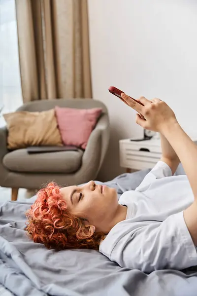Привлекательный расслабляющий странный человек в домашней одежде с рыжими волосами, лежащими на кровати и использующий ее смартфон — стоковое фото