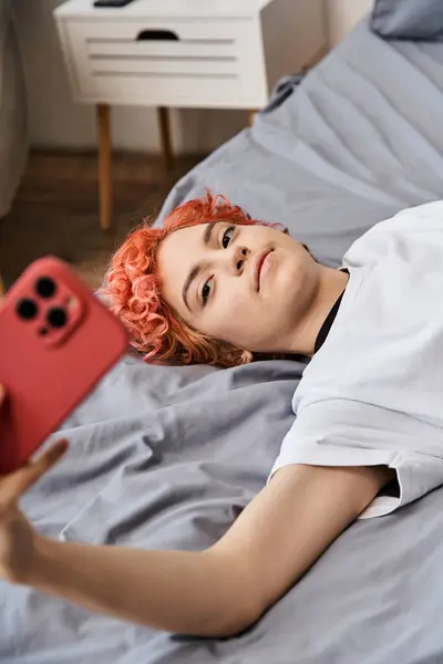 Молодой расслабляющий педик в домашней одежде с рыжими волосами, лежащими на кровати и делающими селфи на телефоне — стоковое фото