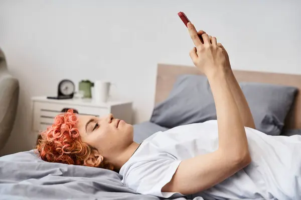 Привлекательный расслабляющий странный человек в домашней одежде с рыжими волосами, лежащими на кровати и использующий ее смартфон — стоковое фото