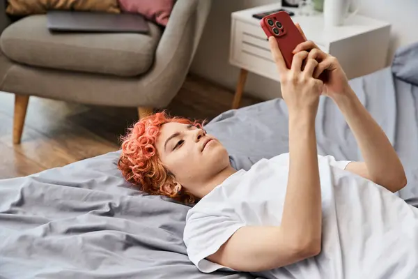 Atraente relaxante queer pessoa em casa com cabelo vermelho deitado na cama e usando seu smartphone — Fotografia de Stock