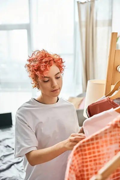 Bela pessoa extravagante de cabelos vermelhos pegando roupas elegantes perto de rack em casa, tempo de lazer — Fotografia de Stock
