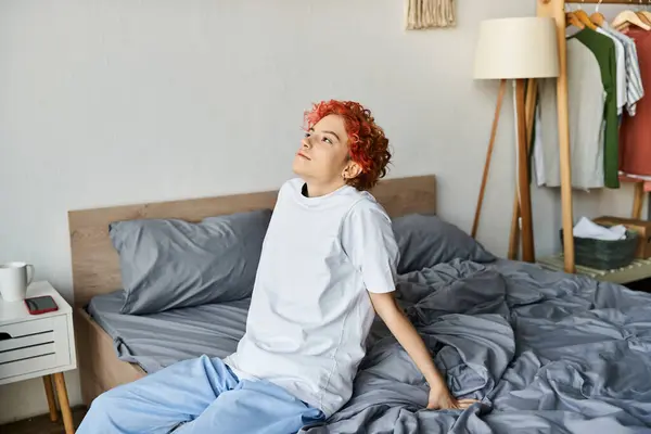 Persona extravagante bien parecido en camiseta blanca con el pelo rojo sentado en su cama, tiempo libre - foto de stock