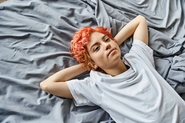 Atractiva persona extravagante con el pelo rojo vibrante acostado en su cama y mirando hacia otro lado, tiempo libre - foto de stock