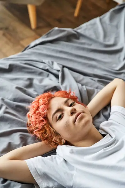 Alegre atraente queer pessoa com cabelos vermelhos deitado em sua cama e olhando para a câmera, tempo de lazer — Fotografia de Stock