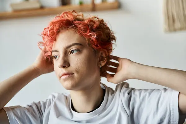 Atractiva persona queer con el pelo rojo vibrante sentado en la cama y mirando hacia otro lado, tiempo libre - foto de stock