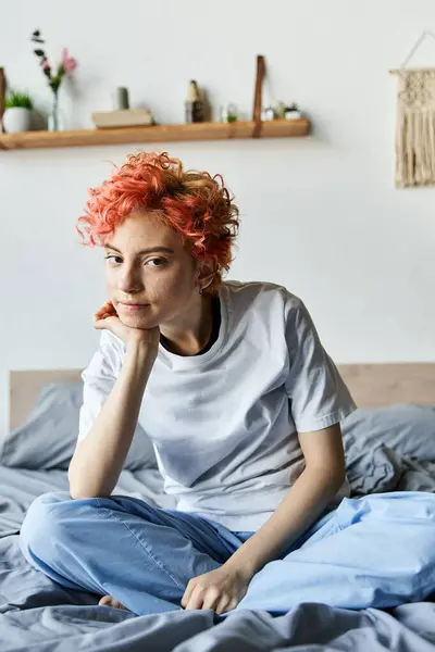 Hermosa persona queer con el pelo rojo vibrante sentado en la cama y mirando a la cámara, tiempo libre - foto de stock