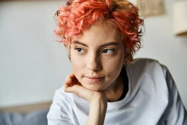 Atractiva persona queer con el pelo rojo vibrante sentado en la cama y mirando hacia otro lado, tiempo libre - foto de stock
