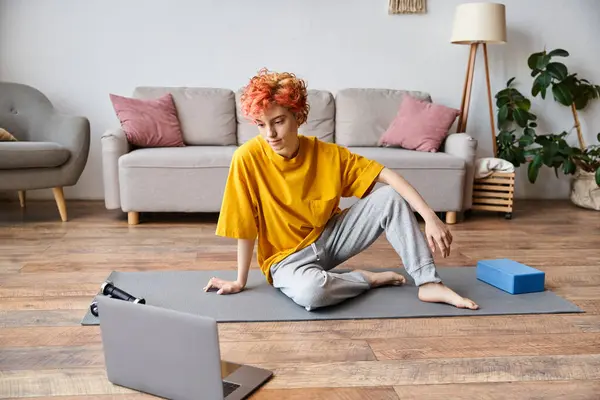 Hermosa persona queer en camiseta amarilla sentado en la esterilla de yoga y mirando a la computadora portátil antes de hacer ejercicio - foto de stock