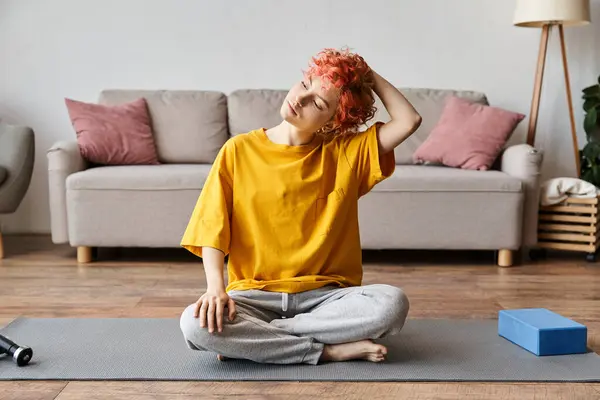 Atractivo joven queer persona en vívida camiseta amarilla sentado en la estera y haciendo un poco de yoga en casa - foto de stock