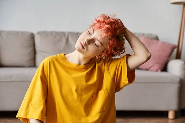 Atractivo joven queer persona en vívida camiseta amarilla sentado en la estera y haciendo un poco de yoga en casa - foto de stock