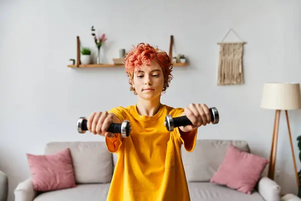 Atractivo joven queer en vibrante traje deportivo ejercicio con mancuernas, mientras que en casa - foto de stock