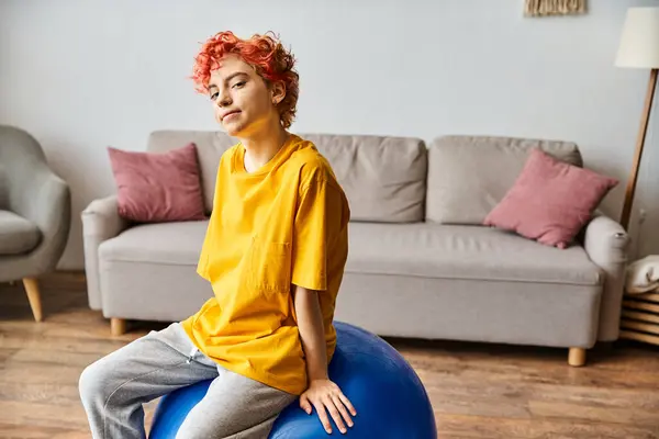 Alegre queer pessoa com cabelos vermelhos sentado na bola de fitness e sorrindo para a câmera enquanto em casa — Fotografia de Stock