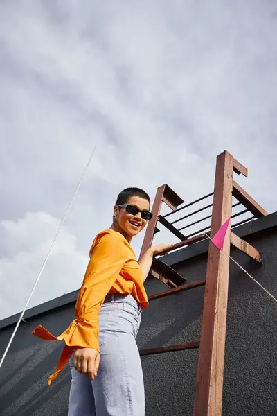 Joyous young caucasian woman in vibrant urban attire posing on stairs on roof and smiling at camera — Stock Photo