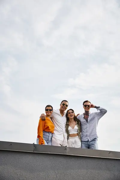 Four diverse jolly friends with sunglasses in vivid attires posing on rooftop and smiling at camera — Stock Photo