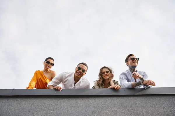 Jolly multicultural friends in urban clothing with sunglasses posing on roof and smiling at camera — Stock Photo