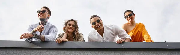 Joyous multiracial friends in urban clothing with sunglasses posing on roof and smiling at camera — Stock Photo