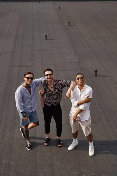 Three diverse cheerful male friends with sunglasses posing on rooftop and looking at camera — Stock Photo