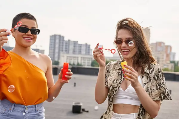 Attraenti donne gioiose in abiti casual vivaci con occhiali da sole che soffiano bolle di sapone sul tetto — Foto stock
