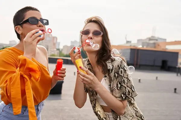 Attraenti giovani donne in abiti casual vivaci con occhiali da sole che soffiano bolle di sapone sul tetto — Foto stock