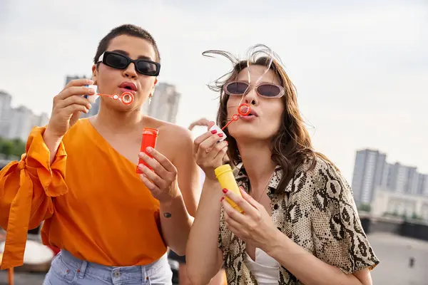 Good looking young women in vibrant casual outfits with sunglasses blowing soap bubbles on rooftop — Stock Photo