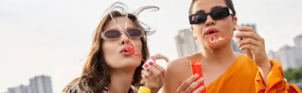 Beautiful jolly women in vibrant outfits with sunglasses blowing soap bubbles on rooftop, banner — Stock Photo