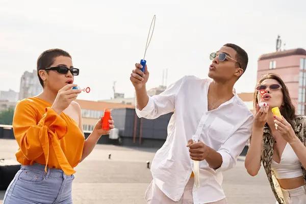 Gioiosi amici multiculturali con eleganti occhiali da sole che soffiano bolle di sapone sul tetto insieme — Foto stock