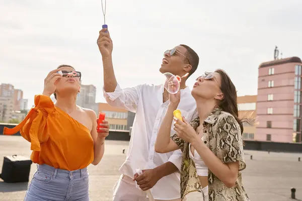 Joyful multiracial friends with stylish sunglasses blowing soap bubbles on rooftop together — Stock Photo