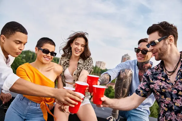 Amigos multirraciales alegres con gafas de sol tintineando sus tazas rojas con bebidas mientras están en la azotea - foto de stock