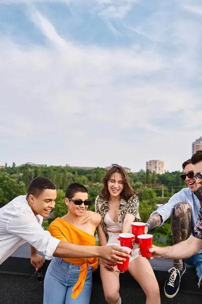 Amis interracial joyeux avec des lunettes de soleil accrochant leurs tasses rouges avec des boissons tout sur le toit — Photo de stock