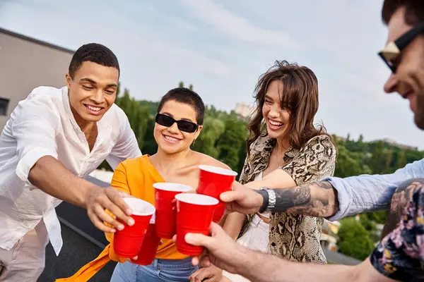 Allegri amici diversi con occhiali da sole clinking loro tazze rosse con bevande mentre sul tetto — Foto stock