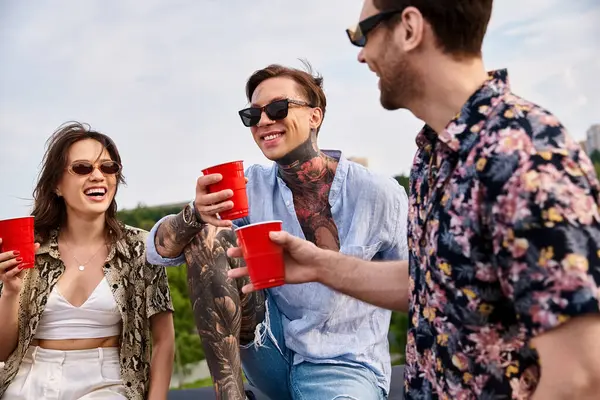 Fröhlich gut aussehende Freunde in lebendigen Klamotten klappern ihre roten Tassen mit Getränken auf dem Dach — Stockfoto