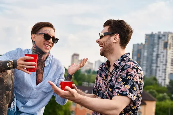 Dos hombres alegres en traje casual con gafas de sol elegantes hablando y sosteniendo copas rojas con bebidas - foto de stock