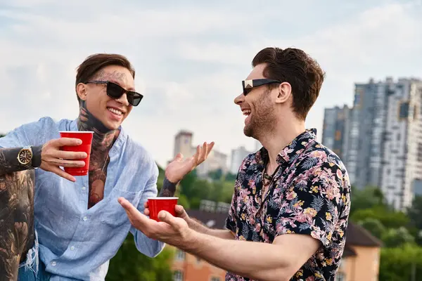 Dos hombres alegres en traje casual con gafas de sol elegantes hablando y sosteniendo copas rojas con bebidas - foto de stock