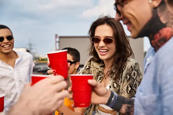 Alegre atractivo interracial amigos divertirse en la fiesta en la azotea y tazas rojas tintineo con bebidas - foto de stock
