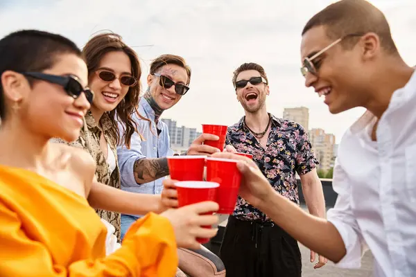 Веселые разнообразные друзья в солнечных очках веселятся на вечеринке на крыше, держа красные чашки с напитками — стоковое фото