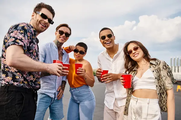 Alegres amigos inter-raciais em roupas vibrantes segurando copos vermelhos com bebidas e sorrindo para a câmera — Fotografia de Stock