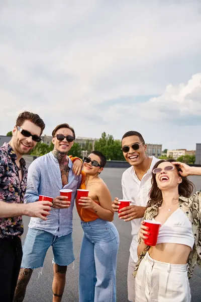 Amigos multirraciales alegres en ropa vibrante sosteniendo copas rojas con bebidas y sonriendo a la cámara - foto de stock