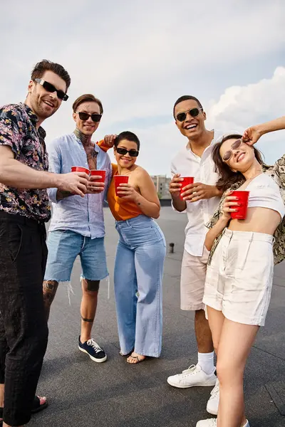 Amigos multiculturais alegres em roupas vibrantes segurando copos vermelhos com bebidas e sorrindo para a câmera — Fotografia de Stock