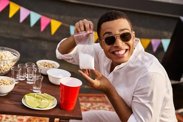 Joyous young african american man holding bag of salt before drinking tequila and smiling at camera — Stock Photo