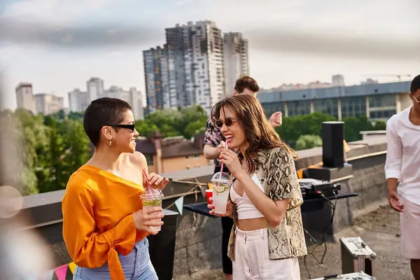 Alegres mujeres atractivas con gafas de sol elegantes beber cócteles junto a sus diversos amigos - foto de stock
