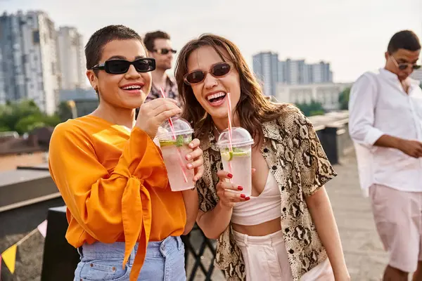 Joyful attractive women with sunglasses drinking cocktails at rooftop party and smiling at camera — Stock Photo