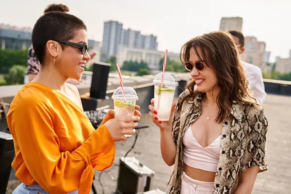 Alegres mujeres guapas con gafas de sol elegantes que beben cócteles junto a sus diversos amigos - foto de stock