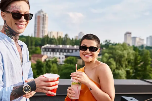 Jolly man and woman in vivid attires with sunglasses drinking at rooftop party and smiling at camera — Stock Photo