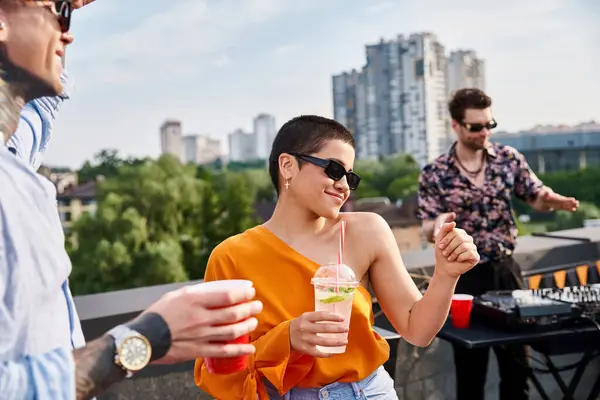 Amigos alegres en atuendos casuales con gafas de sol bebiendo en la fiesta en la azotea y bailando a DJ conjunto - foto de stock
