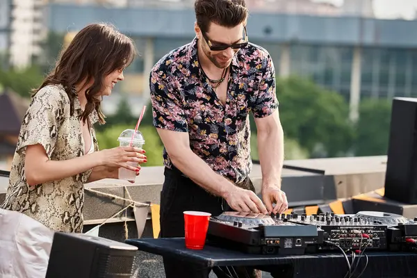 Femme joyeuse avec des lunettes de soleil élégantes et cocktail à la main posant à côté de DJ à la fête sur le toit — Photo de stock