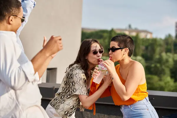 Fröhliche Frauen trinken Cocktail aus Plastikbecher neben afrikanisch-amerikanischer Freundin auf Dachparty — Stockfoto