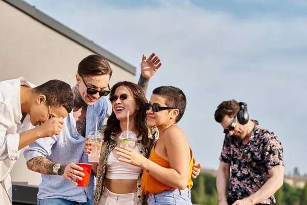 Alegres amigos multiculturales sosteniendo cócteles en las manos y divirtiéndose junto al DJ en la fiesta en la azotea - foto de stock