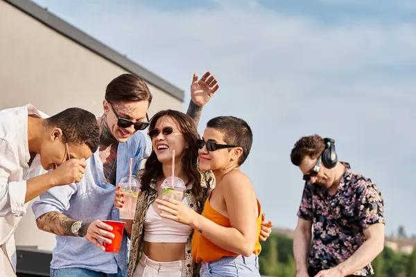 Joyful multicultural friends holding cocktails in hands and having fun next to DJ at rooftop party — Stock Photo