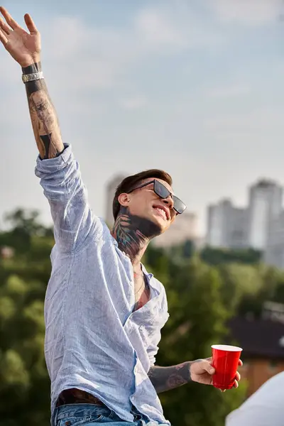 Handsome joyful man with tattoos and sunglasses drinking from red cup and having fun at party — Stock Photo