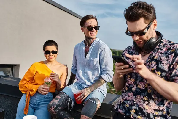 Gutaussehender DJ mit Sonnenbrille und Cocktails auf dem Dach neben seinen fröhlichen Freunden auf sein Handy blickend — Stockfoto